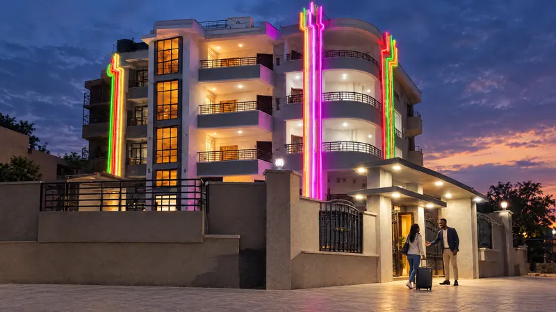 A host welcomes a traveller with a suitcase in front of the lit building at dusk.